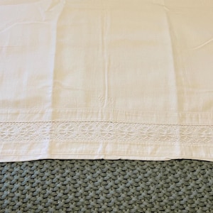 May include: Off-white pillowcase with a decorative floral lace trim along the bottom edge. The pillowcase is made of a smooth, woven fabric and is laying on a textured, teal-colored surface.