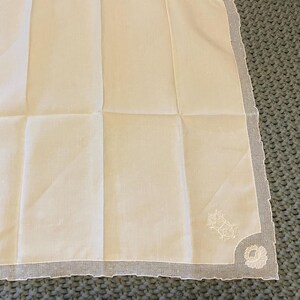 May include: A square, off-white handkerchief with a delicate, scalloped edge. The handkerchief features a floral embroidery design in one corner. The fabric is folded into a grid pattern, suggesting it is new or unused.