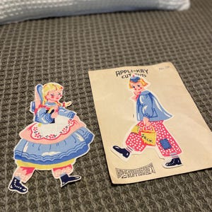 May include: Two vintage paper cutouts of children. One is a girl in a blue dress and pink apron. The other is a boy in a blue jacket and pink polka dot pants. The boy is carrying a bag that says "Jack".