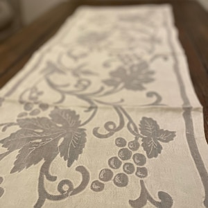 May include: Cream-colored table runner with a gray grape and leaf design. The runner features a repeating pattern of grape clusters and leaves, with a border along the edges. The fabric appears to be linen or a similar material.