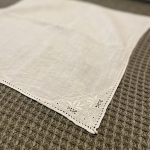 Puede incluir: Un pañuelo blanco cuadrado con detalles bordados florales y geométricos en las esquinas. Los bordes están acabados con un delicado diseño calado. El pañuelo está hecho de una tela suave, posiblemente lino.