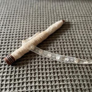 May include: A vintage spool of cream-colored lace ribbon. The spool is wrapped in a matching fabric and has a dark wooden end. The delicate lace features a repeating floral pattern. A classic craft supply.