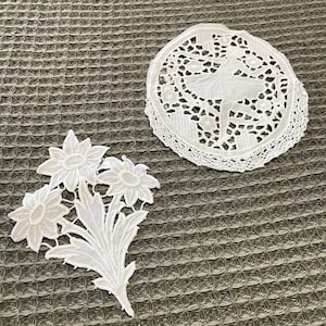 Puede incluir: Dos tapetes de encaje blanco y una aplicación floral se muestran sobre una superficie gris texturizada. Un tapete es redondo con un diseño de bailarina. El otro es una aplicación floral con tres flores y hojas.