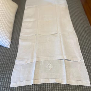 May include: A white linen runner with an embroidered monogram. The runner is rectangular and features a repeating pattern and a decorative border. The monogram is in the center of the runner. The runner is displayed on a gray bedspread.