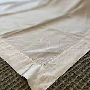 May include: Off-white linen tablecloth with a stitched border and a subtle embroidered initial. The tablecloth is folded, showing a corner with a detailed hem. The fabric has a slightly textured appearance.