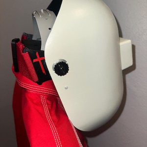 May include: A white welding helmet with a black interior and a black adjustment knob. The helmet is positioned over a red garment with white stitching and a black accent with a red cross.
