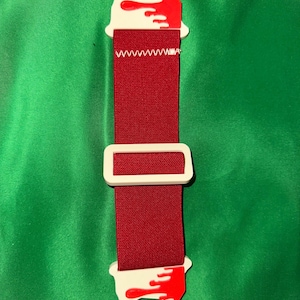 Pode incluir: Um acess&oacute;rio vermelho e branco com uma fivela retangular e duas extremidades circulares. As extremidades brancas t&ecirc;m desenhos de respingos de tinta vermelha. A correia vermelha &eacute; costurada com linha branca.