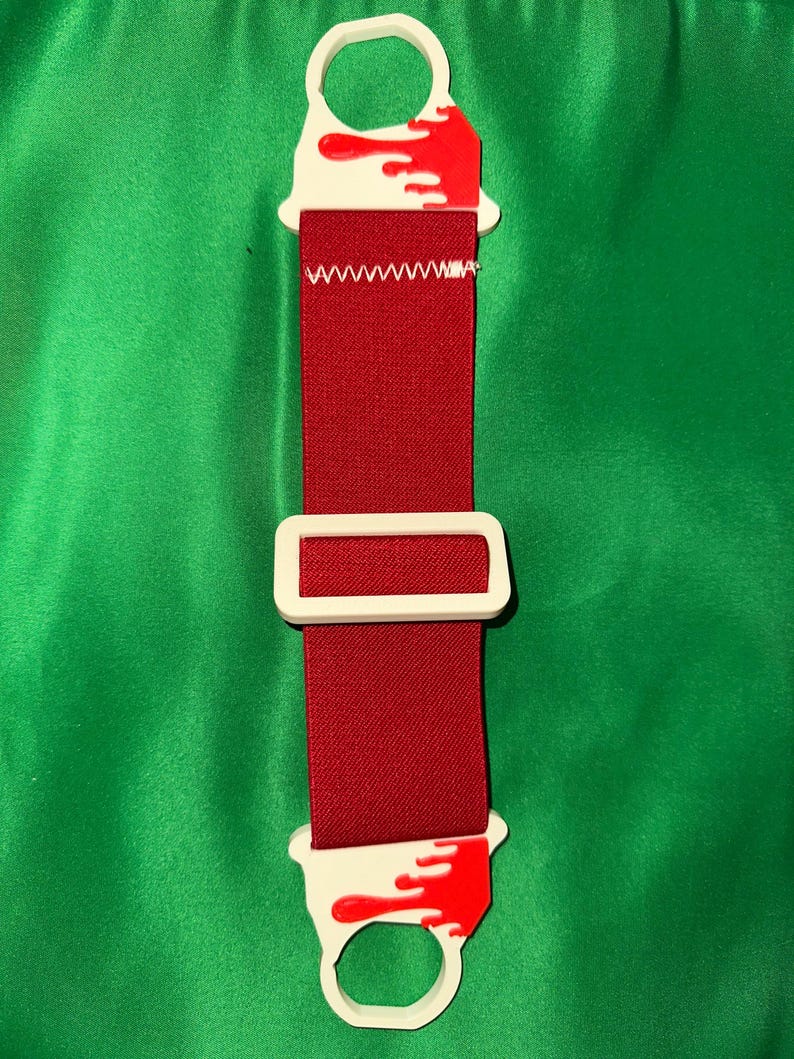 Pode incluir: Um acess&oacute;rio vermelho e branco com uma fivela retangular e duas extremidades circulares. As extremidades brancas t&ecirc;m desenhos de respingos de tinta vermelha. A correia vermelha &eacute; costurada com linha branca.