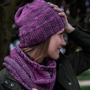 Könnte beinhalten: Ein Strickmützen- und Schal-Set in Lila- und Rosatönen. Die Mütze ist im Oversize-Stil gehalten, und der Schal hat ein Rippenmuster. Das Set ist aus weichem, strukturiertem Garn gefertigt, perfekt für kaltes Wetter. Ein gemütliches und stilvolles Accessoire.