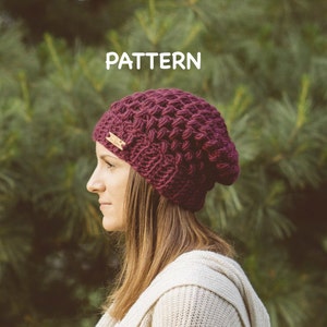 May include: A close-up of a woman wearing a burgundy crocheted beanie with a small wooden tag that says "PATTERN". The beanie is textured and has a slouchy fit. The woman is wearing a white sweater and is standing in front of a green background.