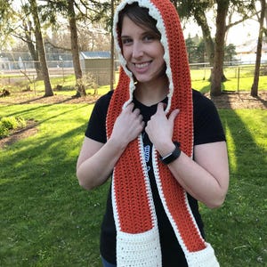 May include: A crocheted scarf with a hood in orange and white. The scarf has two pockets on the ends.