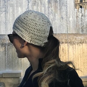 May include: A woman wearing a white crocheted beanie with a ponytail. She is wearing sunglasses and a black jacket.