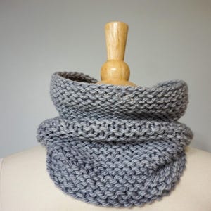 May include: A gray knitted cowl neck scarf, folded and displayed on a wooden mannequin.