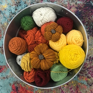Puede incluir: Un cuenco de metal lleno de ovillos de lana en tonos verdes, amarillos, naranjas, marrones y blancos. La lana está dispuesta en forma circular, con el centro del cuenco vacío.