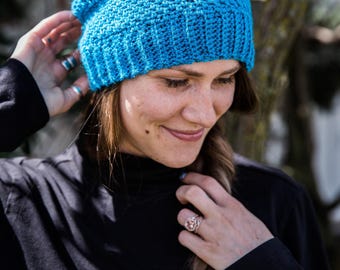 May include: A woman wearing a blue crocheted beanie with a textured pattern. She is wearing a black mock neck shirt and has long brown hair.