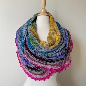 Pode incluir: Um xale de crochê colorido com uma borda de crochê rosa. O xale está drapeado sobre um manequim branco.