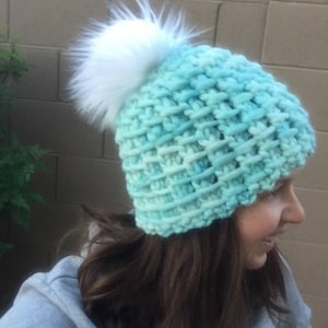 May include: A teal blue knitted beanie with a white faux fur pom pom. The beanie is made with a chunky yarn and has a textured stitch pattern.