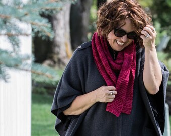 May include: A woman wearing a dark gray poncho with a red knitted scarf. She is smiling and wearing sunglasses.