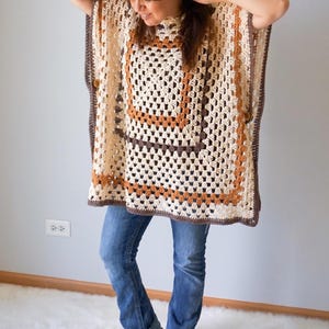 Op de afbeelding: Een gebreid poncho met een geometrisch patroon in bruin, oranje en wit. De poncho heeft een hoge hals en een losse pasvorm.