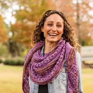 May include: A woman wearing a purple and brown knitted shawl over a denim jacket. The shawl has a lace-like pattern. The background features autumn foliage and a grassy area, creating a warm, natural setting.