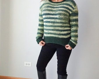 Puede incluir: Un suéter de crochet a rayas verdes y blancas con mangas largas. El suéter se lleva con jeans negros y botas negras de caña alta.
