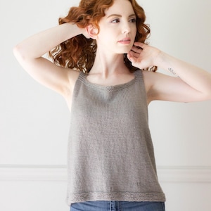 Op de afbeelding: Een vrouw met rood krullend haar draagt een grijs, mouwloos gebreid tanktop en een blauwe jeans.