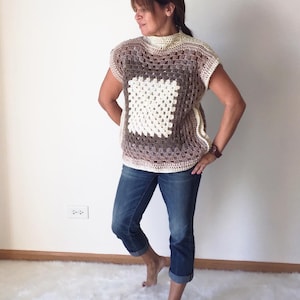May include: A crocheted top with a brown, white, and beige color gradient. The top has a square pattern in the center and short sleeves.
