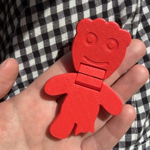 May include: A bright red, articulated figure shaped like a gummy candy, featuring a rounded head with embossed circular eyes and a curved mouth, a rectangular neck segment, and a body with outstretched arms and legs, one of which appears bitten.