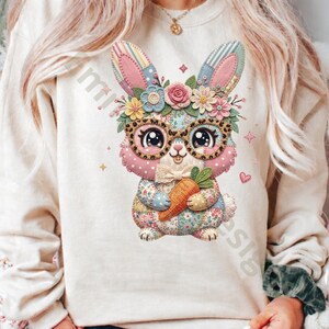 May include: A cream-colored sweatshirt featuring a colorful, cartoon-style bunny design. The bunny wears glasses and a floral crown, holding a carrot. The design incorporates pink, blue, yellow, and orange hues, with a patchwork aesthetic.