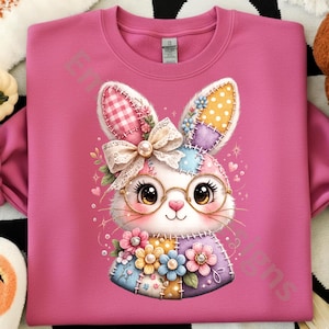 May include: A pink sweatshirt featuring a colorful, patchwork-style bunny wearing glasses and a bow. The bunny has large eyes and is surrounded by flowers and hearts. The design is whimsical and suitable for Easter or spring.