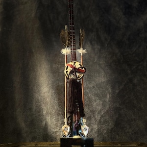 Puede incluir: Un juego decorativo japonés de arco y flechas. El arco es de color marrón oscuro con un emblema circular dorado y rojo. Se adjuntan dos flechas con plumas. Una larga borla marrón oscuro cuelga del arco.