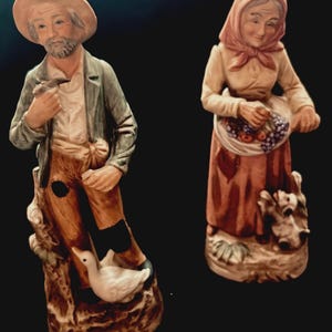 May include: A pair of ceramic figurines depicting an elderly farmer and his wife. The farmer wears a hat, green jacket, and holds a tool. His wife wears a headscarf and apron, carrying a basket of fruit. The figurines are painted in muted tones.