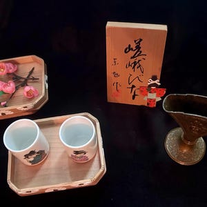 May include: An assortment of Japanese decorative items on a black surface. Includes wooden trays with cups and flowers, a wooden box with calligraphy and a doll, and a bronze-colored vase.
