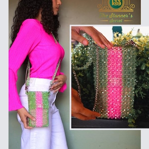 May include: A beaded crossbody phone bag in shades of green and pink, with a chain strap. The bag is held by a person wearing a bright pink long-sleeve shirt and white pants. The bag has a vertical stripe design.