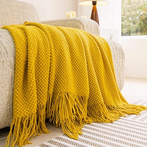 Pode incluir: Cobertor de malha amarelo mostarda com franjas, drapeado sobre um sofá bege. O cobertor tem um padrão texturizado. As franjas pendem sobre um tapete estampado numa sala de estar.