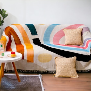 Puede incluir: Un sofá cubierto con una colorida manta con grandes rayas curvas en naranja, turquesa, rosa y azul. Dos cojines beige de punto descansan sobre el sofá. Una pequeña mesa auxiliar blanca con un postre y una botella.