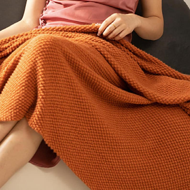 May include: An orange-brown knitted blanket with a textured, raised dot pattern. The blanket is draped over a person's lap, with the edges of the blanket visible. The blanket appears soft and warm, suitable for home decor.