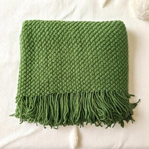 May include: A folded green throw blanket with a textured, bubble-like pattern and a fringe along the bottom edge. The blanket is made of a soft material and is a solid shade of green. The blanket is folded on a white surface.