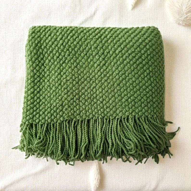 May include: A folded green throw blanket with a textured, bubble-like pattern and a fringe along the bottom edge. The blanket is made of a soft material and is a solid shade of green. The blanket is folded on a white surface.