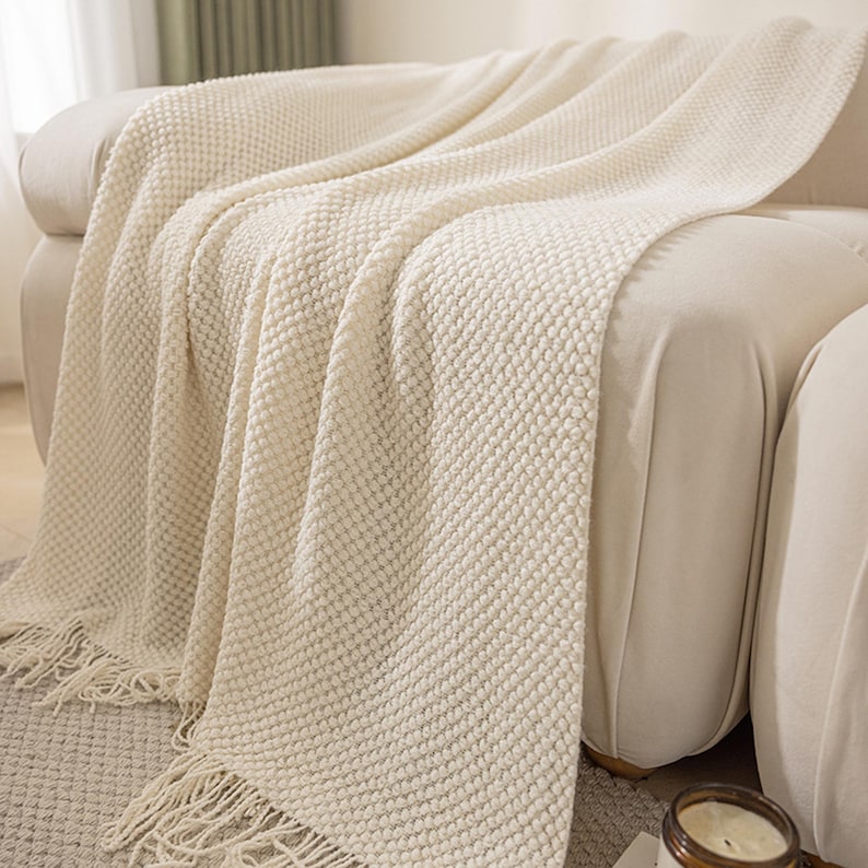 May include: Cream-colored knitted throw blanket with a textured, raised dot pattern. The blanket is draped over a beige sofa, with fringed edges visible. The soft, neutral tones create a cozy and inviting aesthetic.