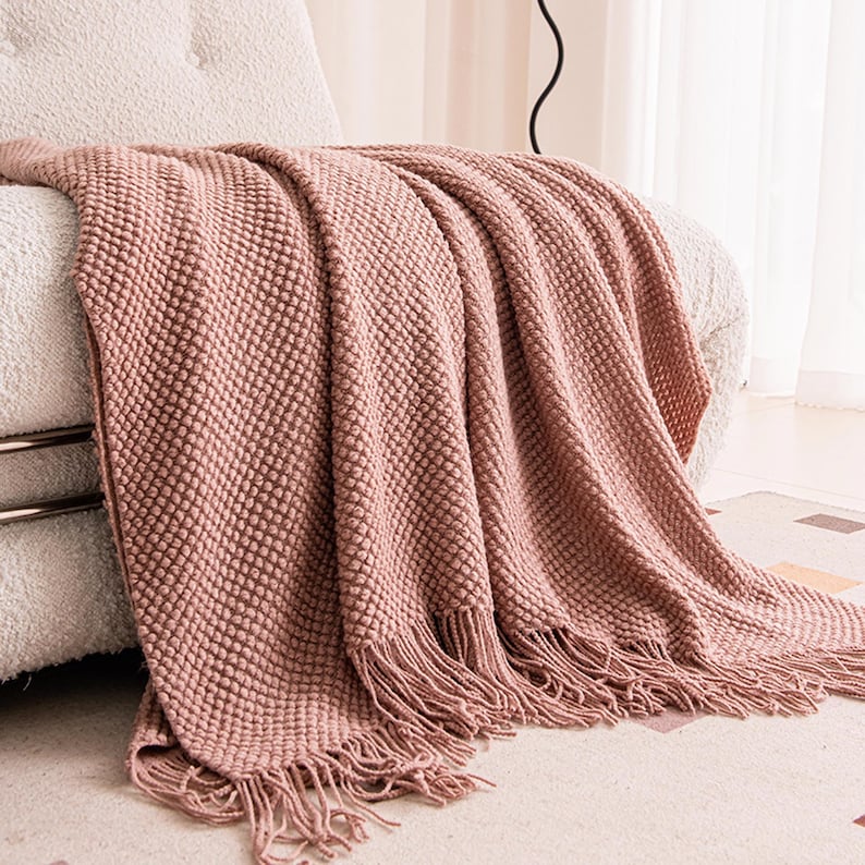 May include: A dusty rose-colored knitted throw blanket with a textured, raised dot pattern. The blanket is draped over a white armchair and features a fringed edge. The soft blanket adds a touch of warmth and texture to any living space.