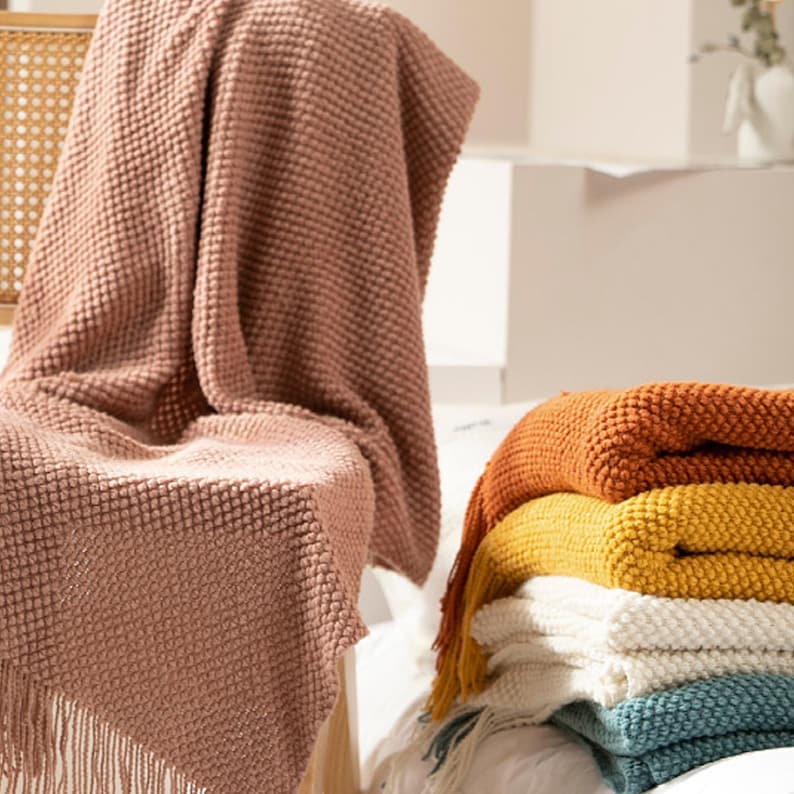 May include: A stack of folded blankets in various colors, including orange, yellow, white, and teal. A dusty rose blanket with fringe is draped over a chair. The blankets have a textured, woven appearance.