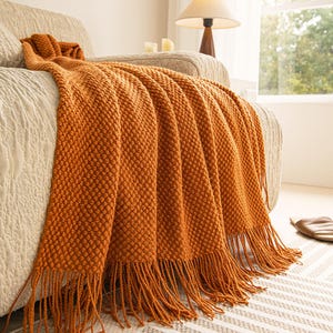 May include: An orange textured throw blanket with fringe detail draped over a beige sofa. The blanket has a woven pattern and is made of a soft material. The throw is a warm, inviting accent for any living space.