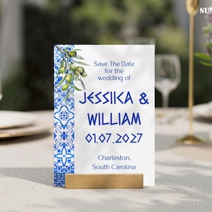 Puede incluir: Una tarjeta de anuncio de boda con un diseño de azulejos azules y blancos y una ilustración de rama de olivo. La tarjeta dice "Save The Date" para Jessica & William, con la fecha 01.07.2027 y la ubicación en Charleston, Carolina del Sur. La tarjeta está en un soporte de madera.