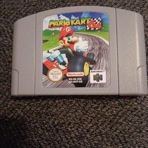 May include: A grey Nintendo 64 video game cartridge for Mario Kart 64. The label features Mario driving a go-kart on a race track with a checkered flag in the background. The text includes Mario Kart 64, Nintendo 64, and the model number NUS-006.