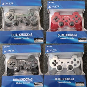 May include: Four Sony PlayStation 3 DualShock 3 wireless controllers in original packaging. The controllers are clear, red, black, and white, each featuring standard directional pads, analog sticks, and action buttons with PlayStation symbols.