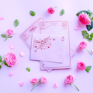 Op de afbeelding: Een stilleven met roze rozen, groene bladeren en verspreide bloemblaadjes. Vintage ansichtkaarten en enveloppen met rode accenten zijn gerangschikt op een lichtpaars oppervlak. De enveloppen hebben een vogel- en bloemenontwerp.