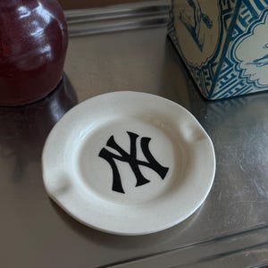 May include: A round, off-white ceramic ashtray featuring a speckled texture and a black New York Yankees logo printed in the center. The ashtray has two notches along the rim for resting items, with a smooth, glossy finish across the surface.