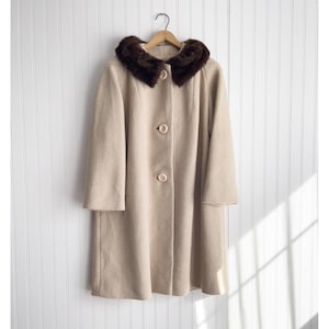 May include: A vintage beige coat with a dark brown fur collar hangs on a wooden hanger. The coat features a button-down front and long sleeves. The background is a white, vertical-slat wall.