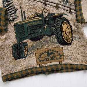 Puede incluir: Un su&eacute;ter beige con un dise&ntilde;o tejido verde y marr&oacute;n de un tractor John Deere. El su&eacute;ter tiene un ribete de cuadros verde y marr&oacute;n alrededor de los bordes. El tractor tiene el texto "John Deere" y "Moline, Ill." en &eacute;l.
